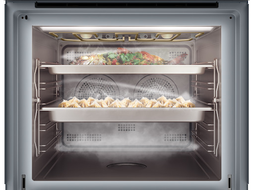 Interior view of a combi-steam oven filled with steam, showing two racks of food cooking simultaneously: a whole fish on the top rack and a tray of dumplings on the bottom rack.
